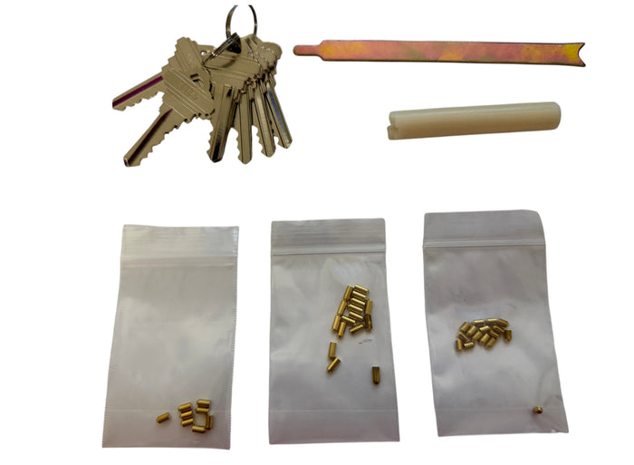 A lock pick kit displayed on a white background, including a set of keys, a metal tool, a cylindrical piece, and three small zippered bags containing brass-colored lock components.