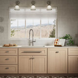 Light wood kitchen cabinets with silver handles, a white sink, and a window with a view of a desert landscape. Three wall-mounted sconces are above the window, and a cookbook and a small plant sit on the counter.